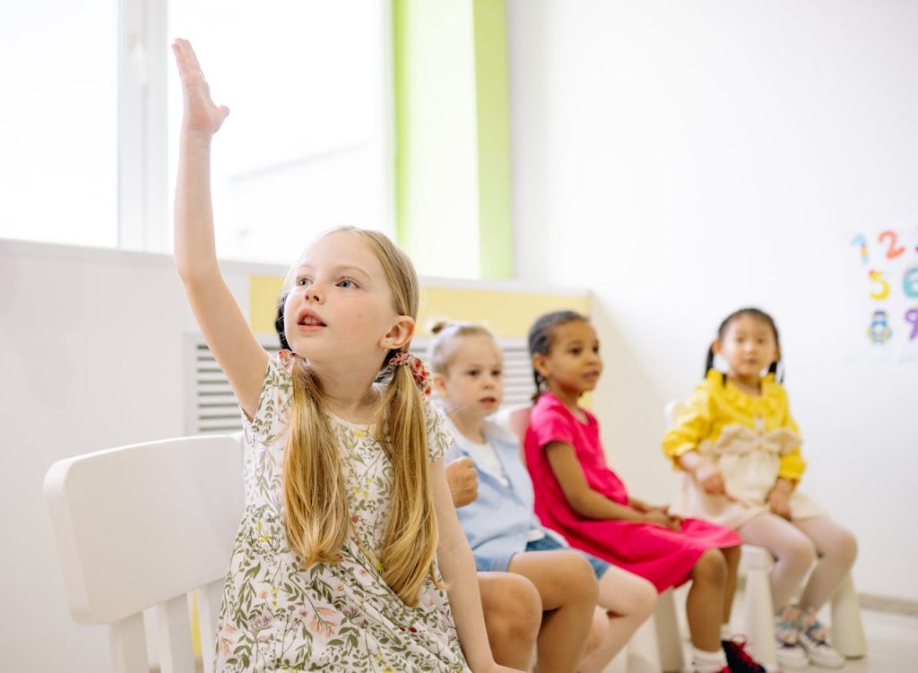 Kindergarten students in a classroom raising hands to participate in learning activities.