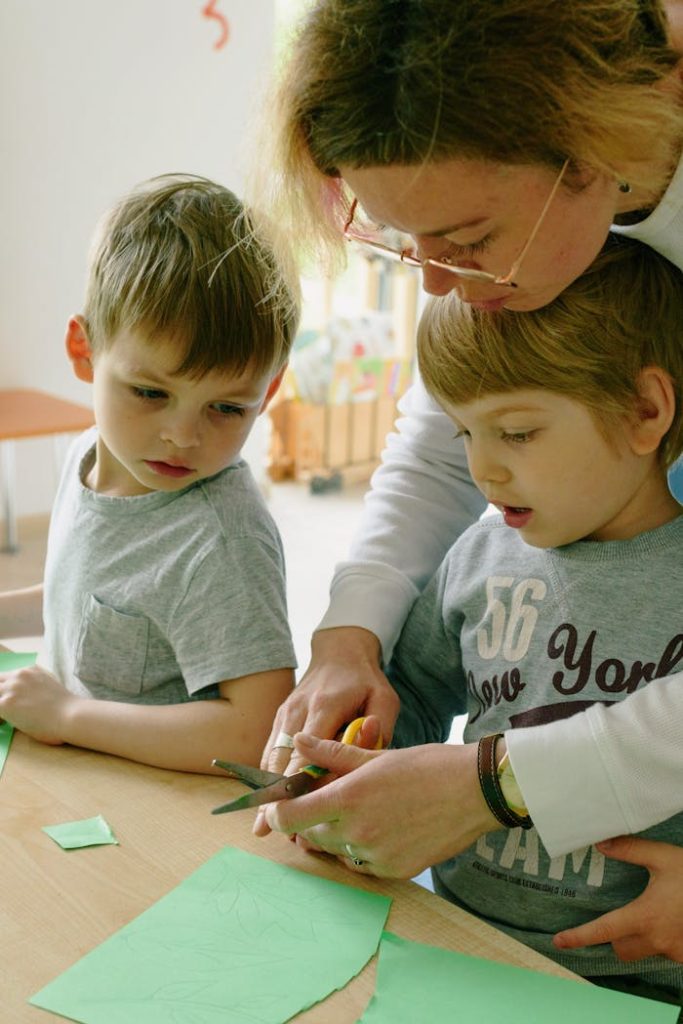 A woman assists two boys with a paper cutting task in a bright classroom setting.